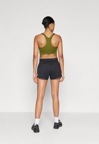 Ελαιόχρωμο αθλητικό σουτιέν με σχέδιο racerback, συνδυασμένο με μαύρα σορτς τρεξίματος. Τα σορτς διαθέτουν ένα διακριτικό μοτίβο και ανακλαστικές λεπτομέρειες.