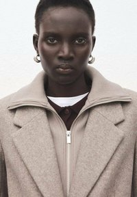 Cappotto beige in misto lana con collo alto e chiusura frontale con zip, indossato sopra un top a coste beige e borgogna. Orecchini a cerchio argento visibili.