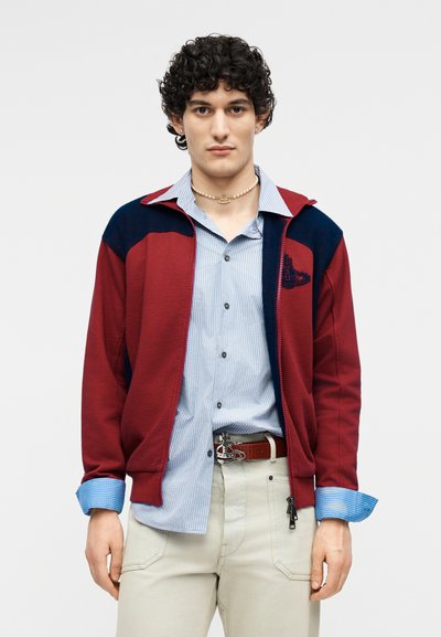 Νέος άνδρας με σγουρά μαλλιά, φοράει κόκκινο και ναυτικό μπουφάν πάνω από μπλε ριγωτό πουκάμισο και μπεζ παντελόνι, στέκεται μπροστά σε απλό φόντο.