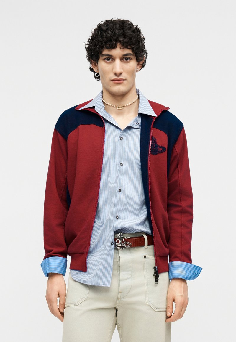 Young man with curly hair wearing a red and navy jacket over a blue striped shirt and beige pants, standing against a plain background.