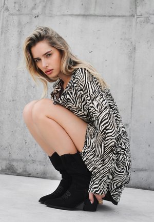 Oversized sukienka koszulowa w czarno-białe paski zebry, z długimi rękawami, w pozycji kucającej, zestawiona z czarnymi zamszowymi botkami za kostkę na słupkowym obcasie.