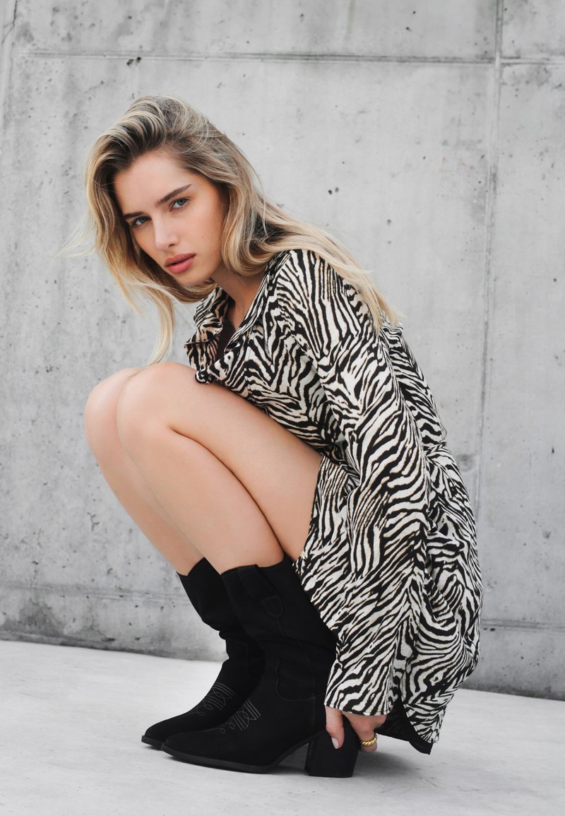 Oversized sukienka koszulowa w czarno-białe paski zebry, z długimi rękawami, w pozycji kucającej, zestawiona z czarnymi zamszowymi botkami za kostkę na słupkowym obcasie.