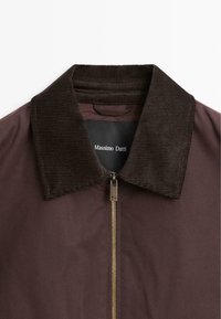 Braune Jacke mit einem Cordkragen und goldenem Reißverschluss. Sie hat eine glatte Textur und ein modernes Design. Das Etikett ist innen am Kragen sichtbar.
