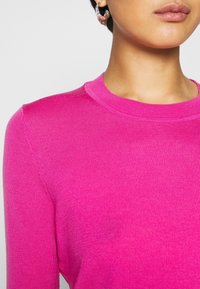 Pull rose ajusté à côtes avec un col rond. Texture lisse et manches longues, avec des détails de couture subtils.