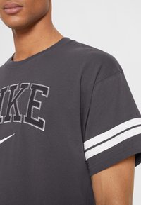 Šedé bavlněné tričko s nápisem "NIKE" ve výrazném stehu a bílými pruhy na rukávech. Měkká textura, klasický střih s kulatým výstřihem.