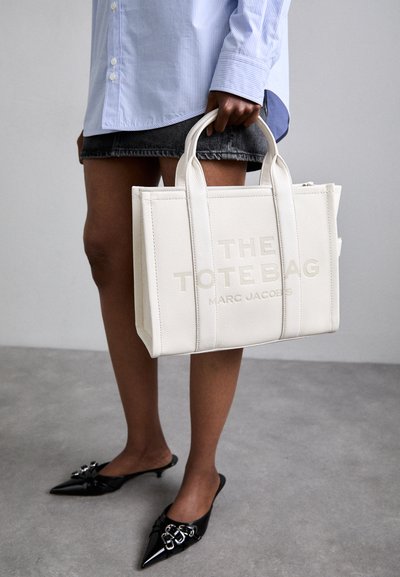 Geantă tote din piele albă cu text embosat "THE TOTE BAG MARC JACOBS." Dispune de mânere doble și o formă structurată, rectangulară.