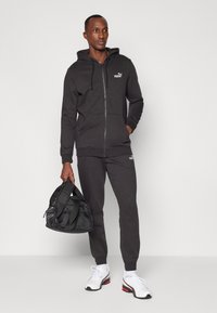 Sort zip-up hoodie og matchende joggingbukser lavet af blødt stof. Har en lille hvid logo. Bærer hvide sneakers og holder en sort taske.