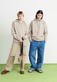 Due persone indossano felpe beige abbinate con un logo. Una abbina la felpa a pantaloni beige strappati, mentre l'altra la indossa con jeans blu larghi e sneakers.