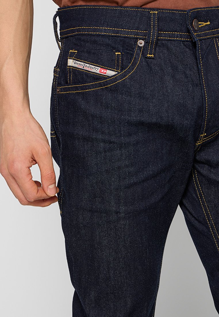 Jeans en denim bleu foncé avec des surpiqûres jaunes, présentant une poche avant avec une étiquette visible. Le matériau semble robuste avec une texture lisse.