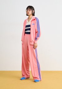 Fato de treino rosa com riscas azuis, casaco com fecho éclair e calças largas. Camiseta listada em branco e azul-marinho por baixo. Calçado azul.