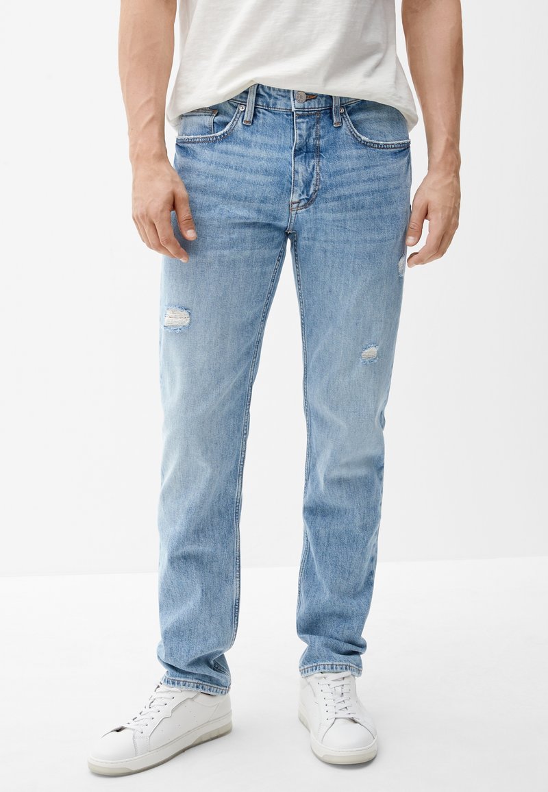 Mann trägt hellblaue, ausgewaschene Jeans, weiße Sneaker und ein weißes T-Shirt, steht vor einem schlichten weißen Hintergrund.