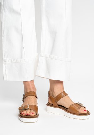 Person wearing white wide-leg pants and brown leather platform sandals with silver buckles and studs, standing on a white floor.