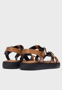 Sandalias de cuero marrón con múltiples diseños de tiras, acentos de hardware dorado y una suela de goma negra. Con la parte trasera abierta y un perfil de suela plana.