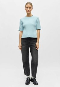 Haut en maille côtelée bleu clair à manches courtes avec bords noirs à festons, associé à un jean noir à jambes droites et des chaussures noires.