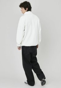 Pull en polaire blanc avec un col montant, associé à un jean noir ample et des chaussures de sport avec des accents gris et noirs.