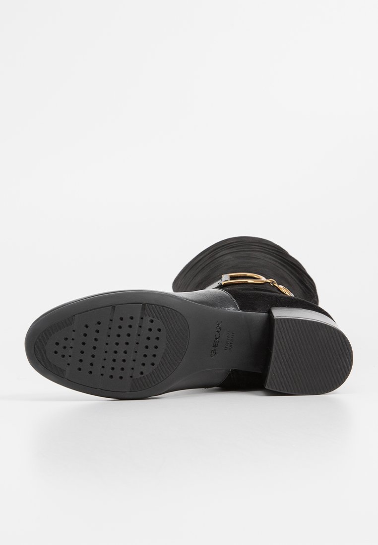 Botte à talon haut en daim noir avec une large sangle, accents en métal doré, surface texturée et semelle en caoutchouc moulé avec de petites perforations.