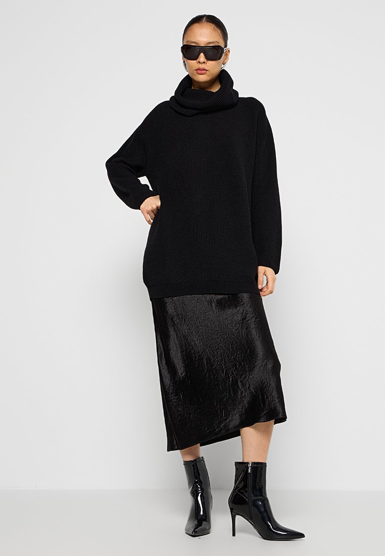 Schwarzer, oversized Strickpullover mit einem Schalkragen, kombiniert mit einem glänzenden, schwarzen Satinstiftrock und schwarzen, hohen Stiefeletten.