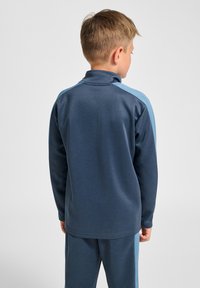 Marineblauwe fleecejas met lichtblauwe paneelaccenten, hoge kraag en zachte textuur. De achterkant heeft een rechte snit en stiksel-details.