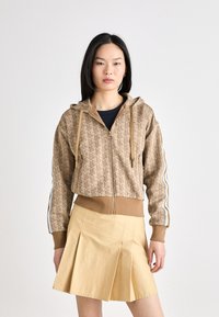 Bruine hoodie met ritssluiting en geometrisch patroon met beige accenten, voorzien van een capuchon en zijstrepen. Vergezeld van een geplooide gele rok.