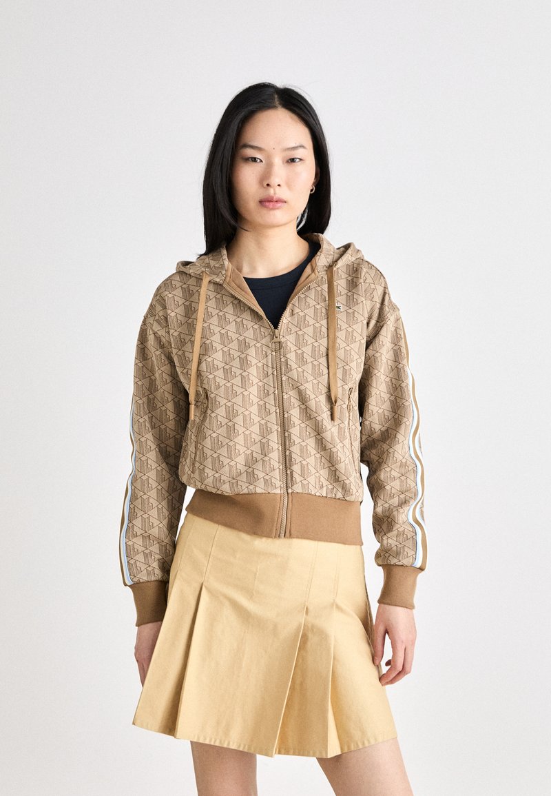 Bruine hoodie met ritssluiting en geometrisch patroon met beige accenten, voorzien van een capuchon en zijstrepen. Vergezeld van een geplooide gele rok.