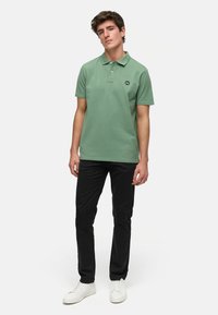 Grön bomullspolo med krage och svart logotyp på bröstet, matchad med svarta byxor och vita sneakers. Enhetlig bakgrund.