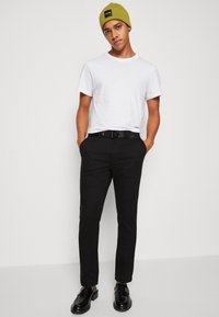 Pantaloni neri a vestibilità slim, abbinati a una maglietta bianca semplice. Un berretto verde e scarpe nere completano l'outfit.