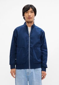 Ναυτικά μπλε ελαφρύ μπουφάν με μπροστινό φερμουάρ, πλάγιες τσέπες και ελαστικούς μανσέτες. Λείο ύφασμα με χαλαρή εφαρμογή.