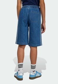 Pantaloncini di jeans con una fascia elastica in vita, di colore blu, lunghezza al ginocchio, con una tasca posteriore e strisce laterali a contrasto. Indossati con scarpe da ginnastica blu.