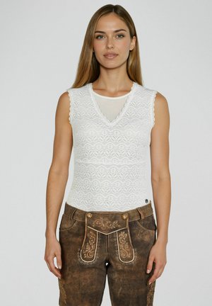 Frau trägt ein weißes, ärmelloses Spitzentop und braune, traditionell bestickte Lederhosen, steht vor einem schlichten Hintergrund.