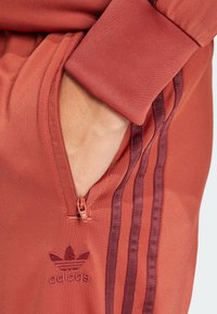 Red athletic pants with a zippered pocket and three vertical maroon stripes. Features an embroidered Adidas logo near the pocket. Texture appears smooth.