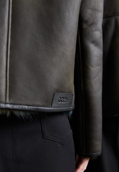 Schwarze Lederjacke mit strukturierter Oberfläche und auffälliger Naht. Besitzt ein kleines Etikett mit geprägten Zahlen, getragen über dunklen Hosen.
