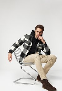 Czarna i biała kraciasta kurtka na czarnej bluzie z kapturem, beżowe spodnie i brązowe buty. Krzesło jest metalowe i wygięte. Tło jest jednolite.