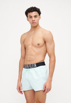 Menta zelene kupaćice s crnim Calvin Klein logom na pojasu, koje imaju podesivu pertlu i glatku, laganu tkaninu.