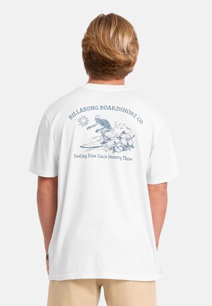 Persona con capelli biondi corti indossa una maglietta bianca con una grafica di uno scheletro che fa surf e il testo "Billabong Boardshort Co" sulla schiena.