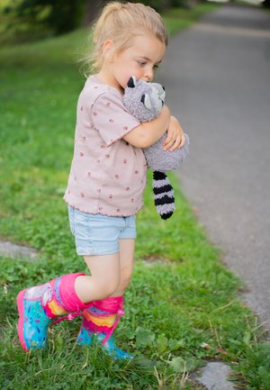 Kind in een lichtroze shirt met kleine bruine stippen, denim shorts en kleurrijke, gestreepte rubberen laarzen, die een grijze raccoon-knuffel omarmt.