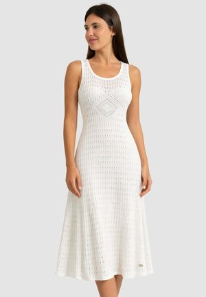 Donna che indossa un abito midi bianco senza maniche, lavorato a maglia con motivo a diamante e linee verticali strutturate, in piedi davanti a uno sfondo semplice.