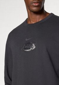 Schwarzes Langarmshirt mit Rundhalsausschnitt, das ein reflektierendes Los Angeles Lakers-Logo und ein kleines Nike-Swoosh zeigt. Aus Baumwollmaterial, weicher Textur.