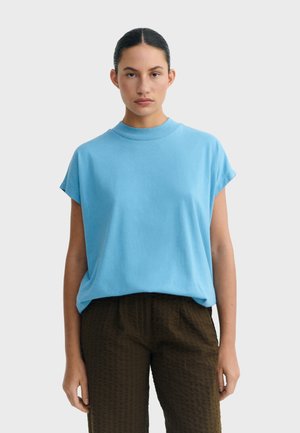 Frau mit zurückgebundenem dunklem Haar, die ein hellblaues kurzärmeliges Oberteil und dunkle, strukturierte Hose trägt und vor einem einfarbigen Hintergrund steht.