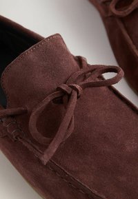 Gros plan sur une chaussure mocassin en daim marron avec des coutures apparentes et un nœud de lacet noué sur le devant.