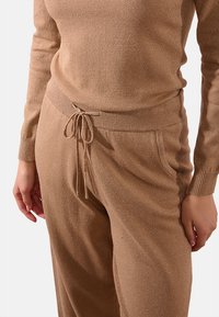 Maglione a maglia color beige e pantaloni jogger abbinati, realizzati in un tessuto leggero e testurizzato con sfumature brillanti. Il maglione ha polsini a coste; i pantaloni jogger presentano un cordoncino in vita e tasche.