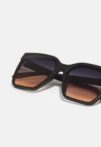 Black square sunglasses with gradient brown lenses, featuring a bold geometric design and thick arms. Lenses transition from dark to light brown.