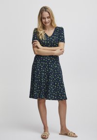 Námořnické šaty s krátkým rukávem, výstřihem do V a vzorem zelených listů. Rozšířený lem, vyrobené z měkké látky, nošené s tělovými sandály.