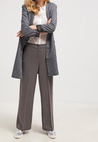 Cardigan long gris porté sur une chemise blanche à boutons avec un motif circulaire, associé à un pantalon gris taille haute et des baskets argentées.
