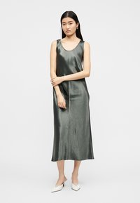 Femeie în picioare purtând o rochie fără mâneci, de culoare gri închis, din mătase și pantofi cu toc ascuțiți albi, pe un fundal simplu și deschis la culoare.