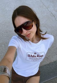 T-shirt blanc avec un graphisme "Rose Blanche" en rouge, associé à un short marron. Grandes lunettes de soleil en écaille de tortue. Bracelet en argent au bras gauche.