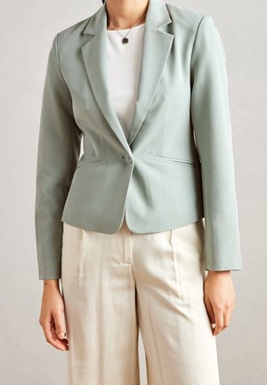 Person wearing a light green tailored blazer with one button over a white shirt and cream-colored pants, against a plain background.