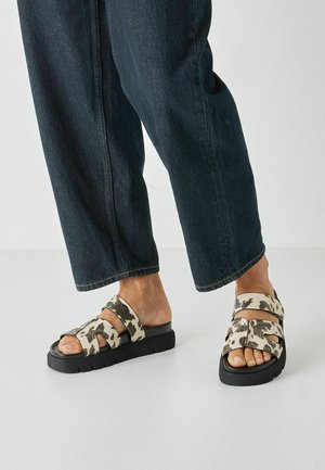 Sandalias negras con suela gruesa, con un diseño de estampado de vaca en las correas. El material parece suave y texturizado. Usadas con jeans oscuros.