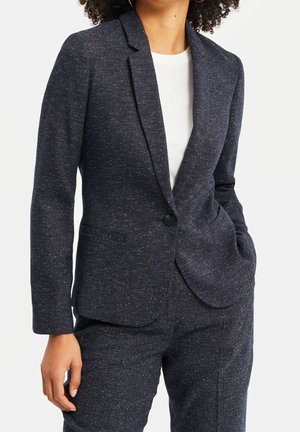 Woman wearing a dark speckled tailored blazer and matching pants with one hand in pocket over a white top, cropped at neck and mid-thigh.