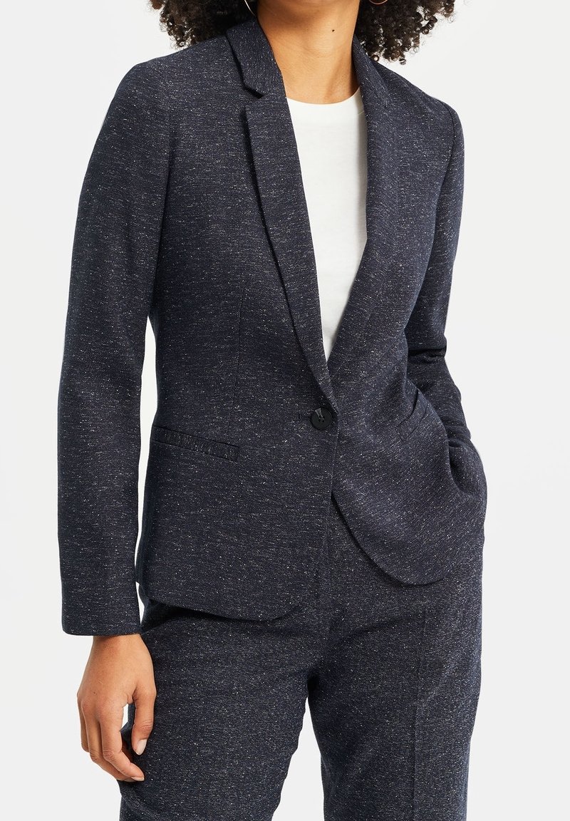 Femme portant un blazer ajusté à pois foncé et un pantalon assorti, une main dans la poche, sur un haut blanc, cadrée au niveau du cou et à mi-cuisse.