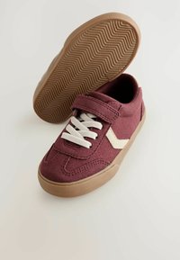 Mørkerøde sneakers med velcro-rem, hvide snørebånd og en struktureret gummisål. Den ene sko står oprejst, hvilket viser indersiden og sålen.